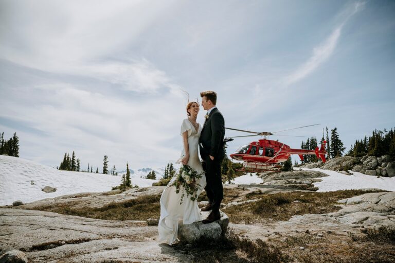A bride and groom holding hands near a red helicopter by Beverley Lake on a rocky mountain landscape with a cloudy sky.