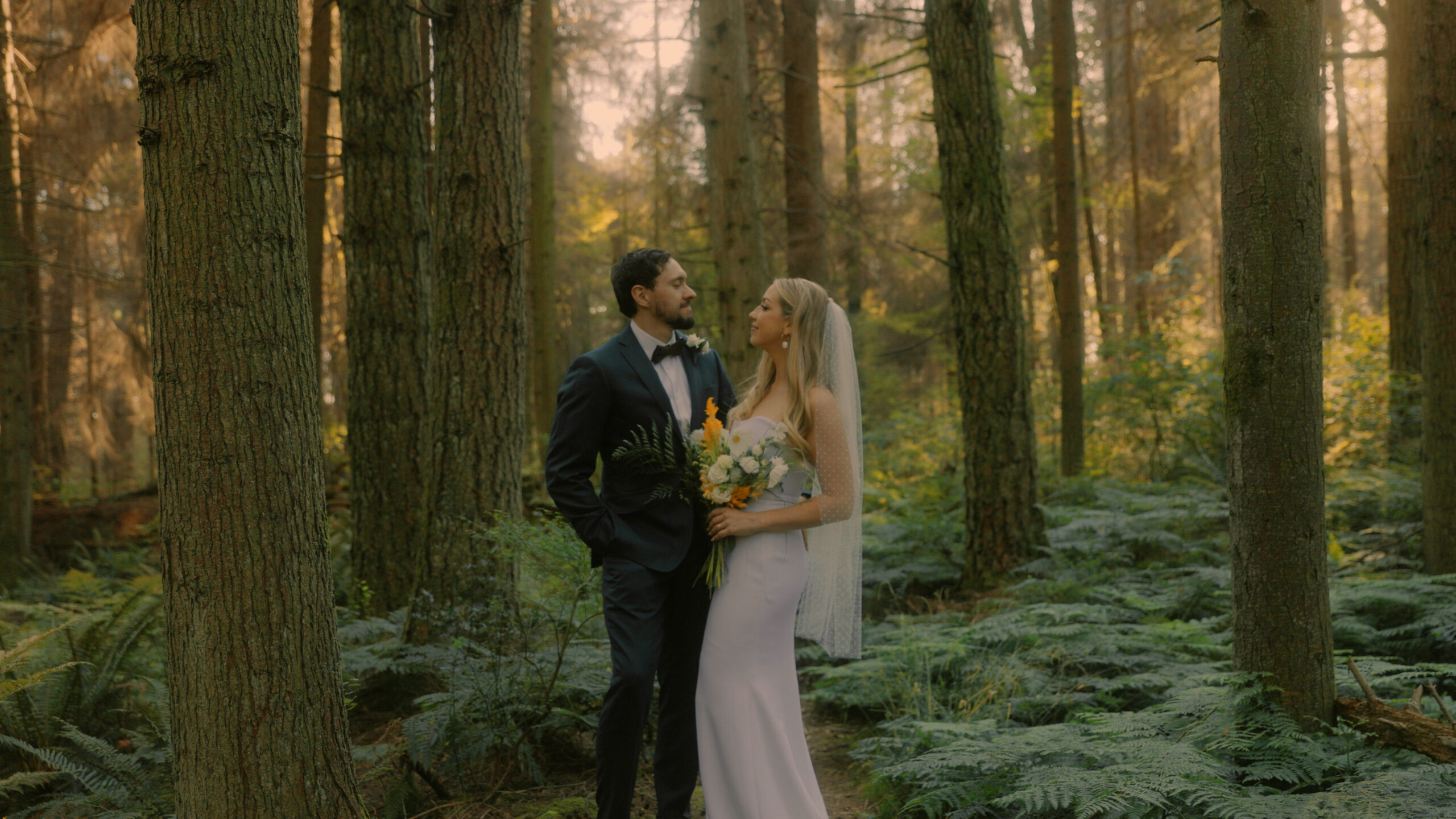 Bride and groom face each other and pose for the camera in the forest at Stanley Park