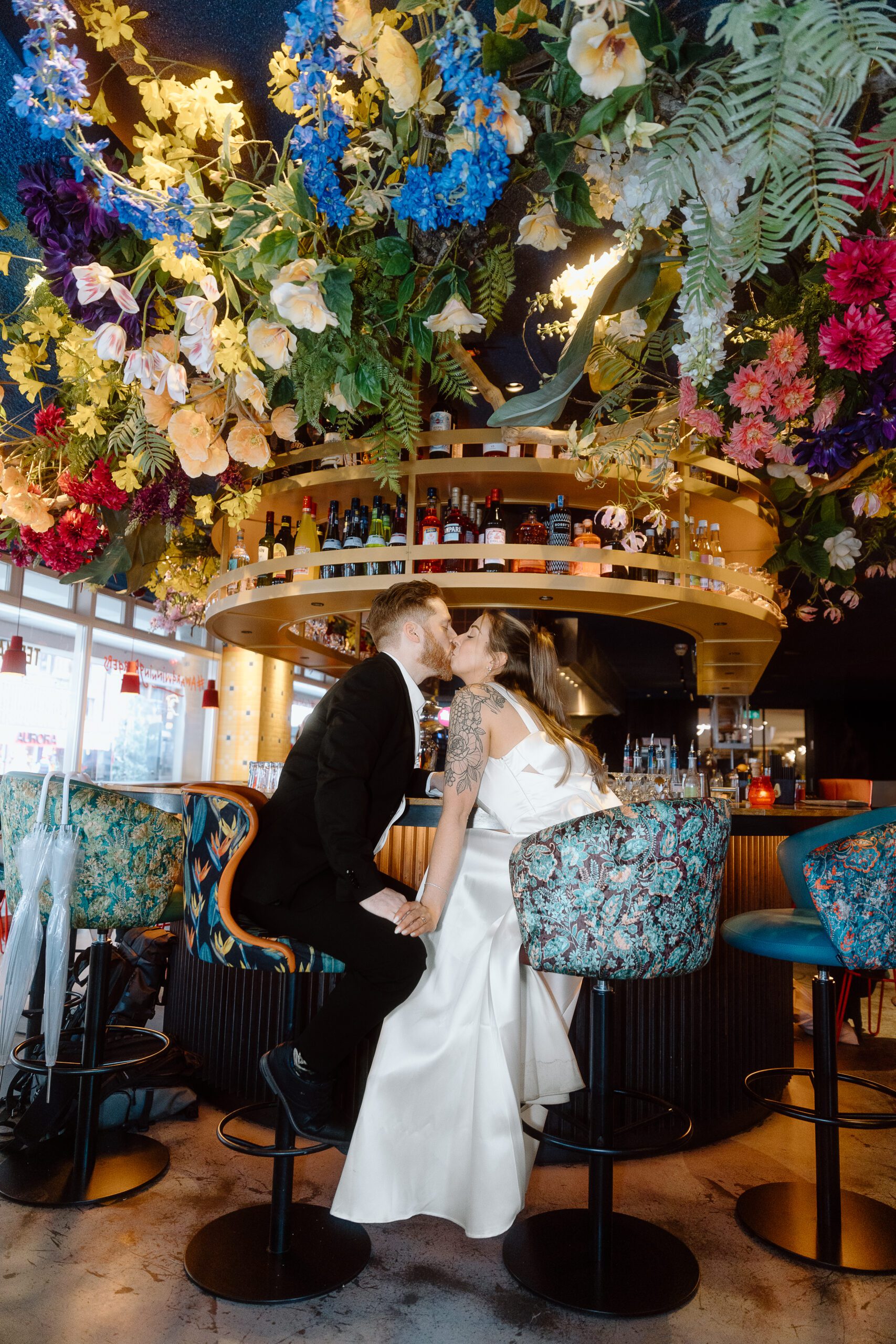 A couple in wedding attire shares a kiss at a bar stool at Ter Marsch & Co in Amsterdam, surrounded by vibrant hanging flowers in a stylish burger bar setting.