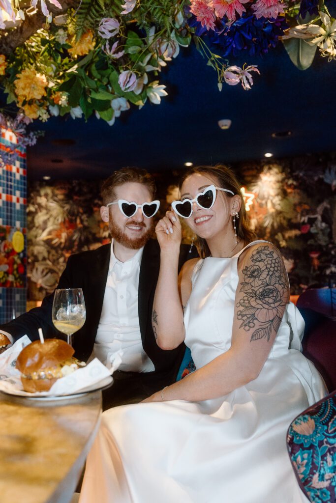 A newlywed couple wearing heart-shaped sunglasses smile while sitting at a bar at Ter Marsch & Co table decorated with flowers in Amsterdam, showcasing a festive and joyful atmosphere.