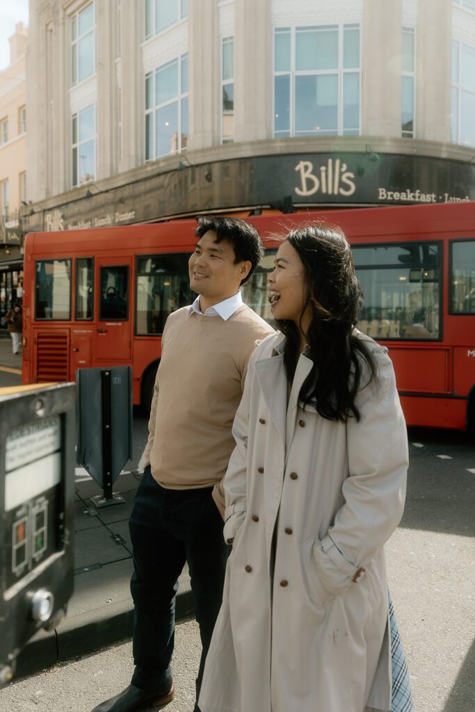 A man and woman smile while waiting at a traffic light in London with a red bus in the background.