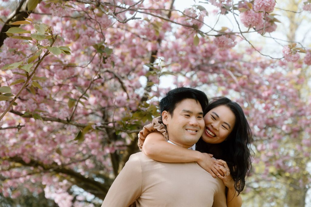 A woman hangs off of a man's shoulders smiling. There are blossom trees in the background.