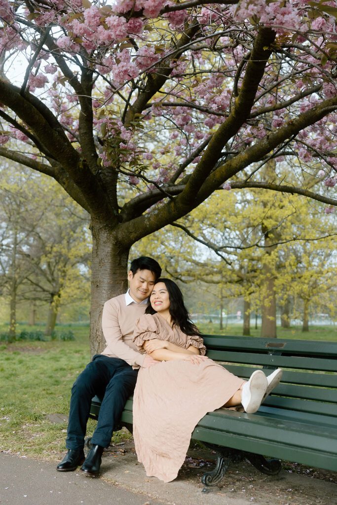 A man and woman cuddle up on a park bench at Greenwich Park in London with blossom tree in the background.