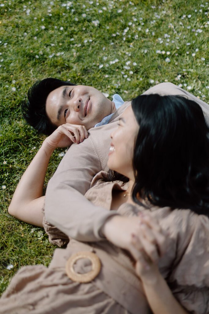 A man and woman lie down in the grass. The woman rests her head on the man's chest.