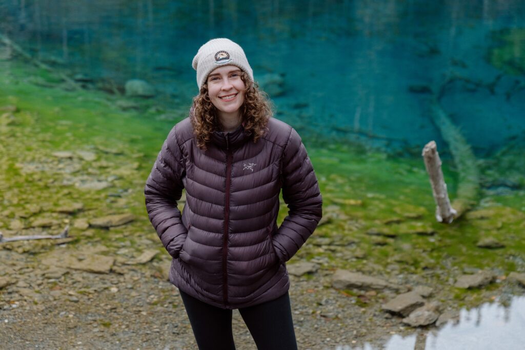 Rachel King, a woman with curly hair, wearing a white beanie and a maroon puffer jacket, smiling in front of a serene, teal-colored lake in Canmore, Alberta.