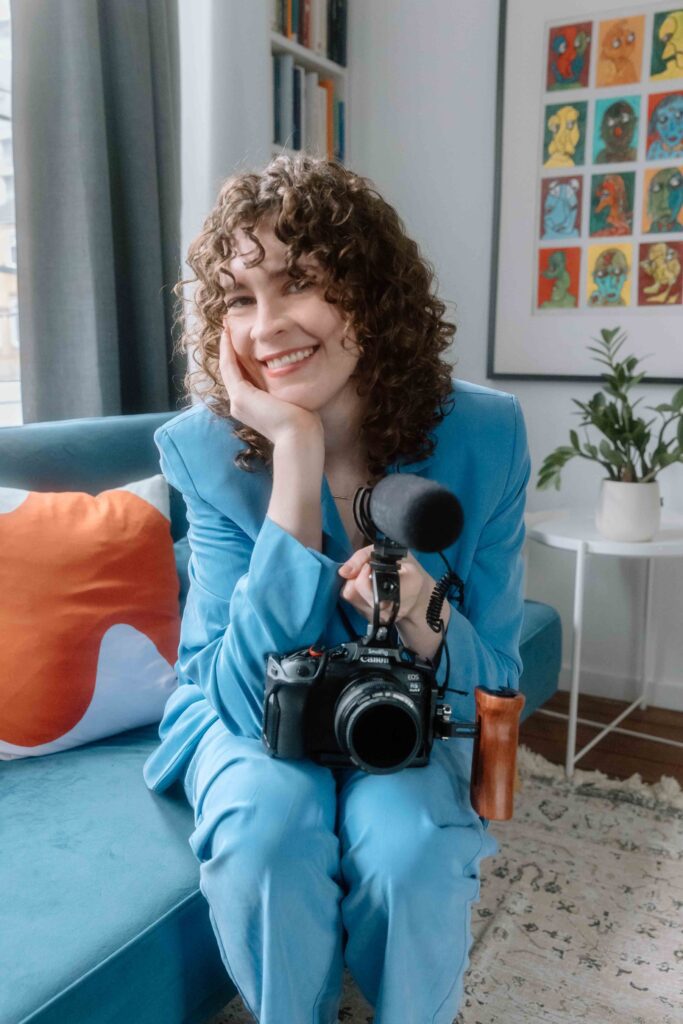 A joyful woman, Rachel King, with curly hair, wearing a blue suit, sits on a couch holding a camera with a microphone attachment, surrounded by vibrant decor and artwork.