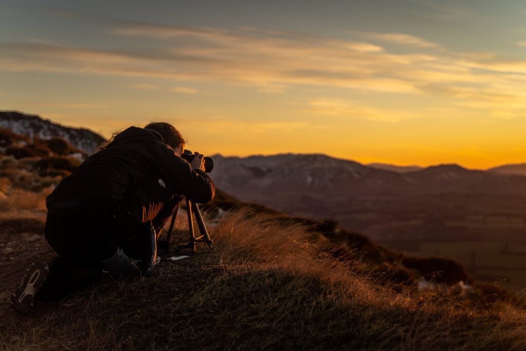 Rachel King crouches, taking pictures during sunset with a camera on a tripod, overlooking a mountainous landscape under a glowing orange sky in New Zealand.