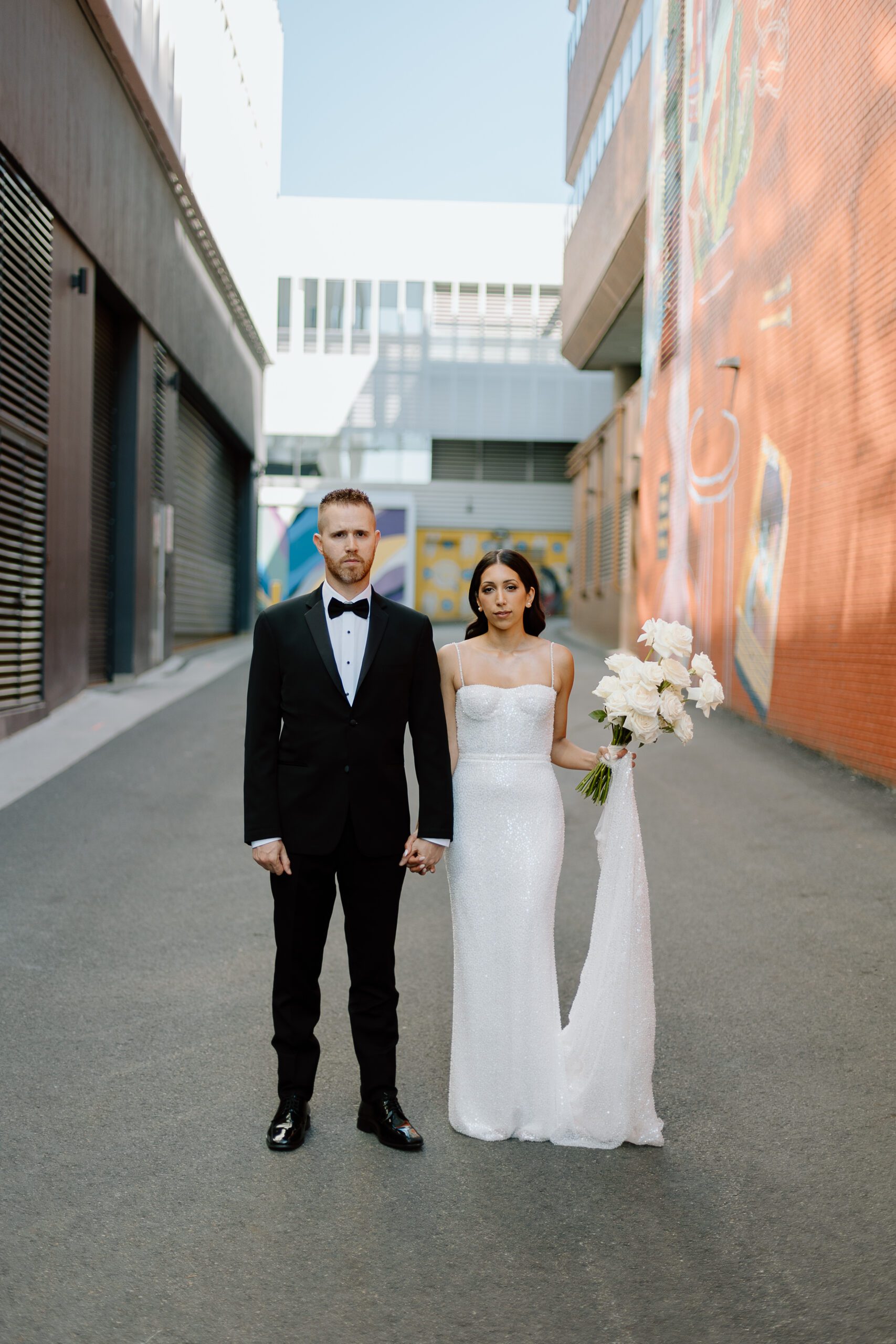 A bride and groom stand holding hands in a Vancouver alleyway, the bride in a white sequined gown with a bouquet, and the groom in a classic black tuxedo.