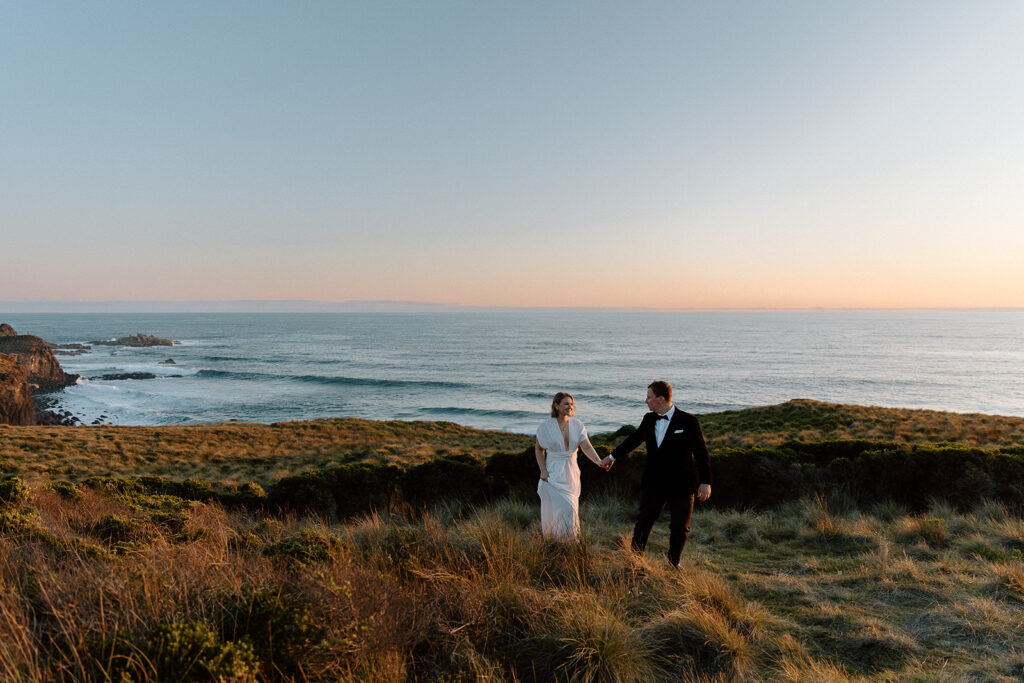 Bride and groom walk along the coast line with the ocean in the background at sunset at Phillip Island.